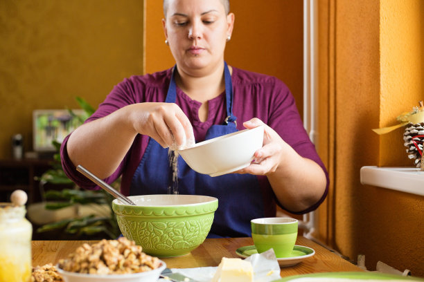 近距离的照片年轻美丽的成年妇女短发，坐在厨房桌子后面，筛面粉到碗，家庭烘焙的概念图片下载