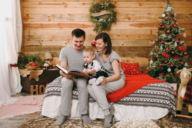 年轻快乐的父母和可爱的孩子在圣诞树旁的床上读童话书在家里装饰的新年木制房间。2018年的生活方式、家庭和度假理念图片下载