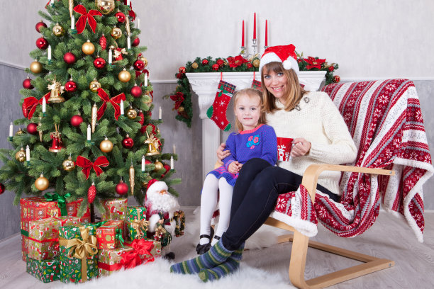 庆祝圣诞节或新年。快乐的母女坐在圣诞树旁的椅子上，拿着圣诞礼物。壁炉的背景是圣诞袜图片下载
