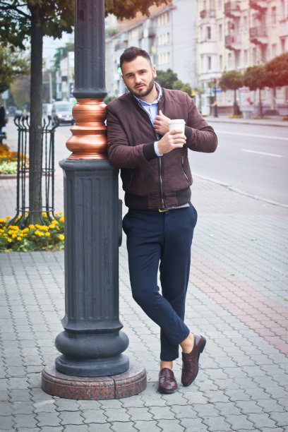 一个穿着夹克和西装的男人走在欧洲的大街上，手里拿着咖啡。商人时尚服装。秋天的风景、肖像。库存图片图片下载