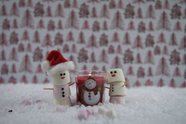 圣诞照片:温暖的红色蜡烛和有趣的棉花糖形状的雪人与冰微笑和圣诞老人帽都站在雪与树图案的背景图片下载