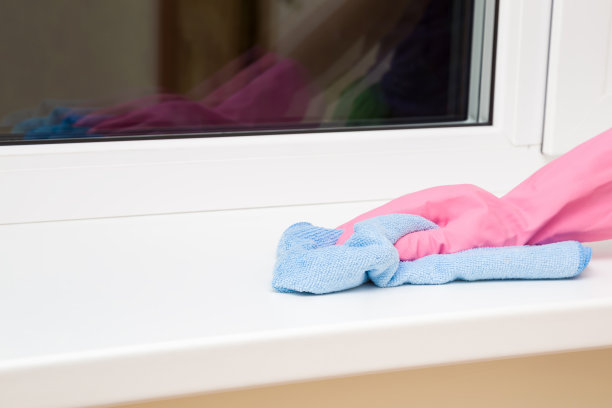 员工手戴橡胶防护手套，用微纤维布擦拭房间窗台上的灰尘。早春或定期大扫除。商业清洁公司理念。素材图片