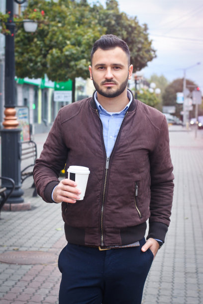一个穿着夹克和西装的男人走在欧洲的大街上，手里拿着咖啡。商人时尚服装。秋天的风景、肖像。库存图片图片下载