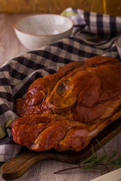 竖着的照片，一大块带骨的烟熏猪肉放在砧板上。蓝色的毛巾、菜刀、叉子、碗和迷迭香，周围是葡萄酒和油，背景是复古墙。图片下载