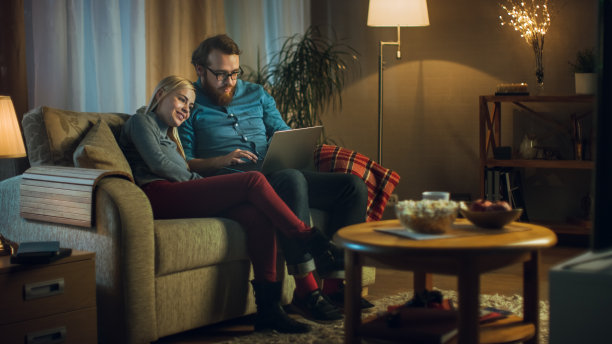 女朋友和男朋友坐在沙发上，膝盖上放着笔记本电脑，看视频，玩得开心。客厅是舒适的。这是晚上。图片下载
