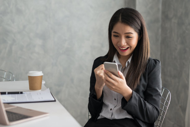兴奋的亚洲商务女孩，而阅读智能手机坐在她的工作站。企业成功的概念图片下载