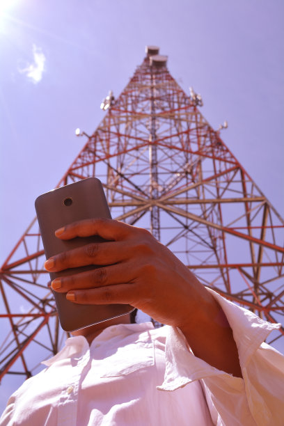 一个拿着智能手机的女人的手。寻找wi - fi连接。在线社交活动和联系概念。非常晴朗的日子。天线，塔移动通信和蓝天背后。图片下载