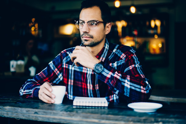 戴着眼镜、戴着时髦手表的胡子青年正在考虑新计划，坐在咖啡厅里享受咖啡休息时间。思考男性与笔和笔记本看向别处图片下载