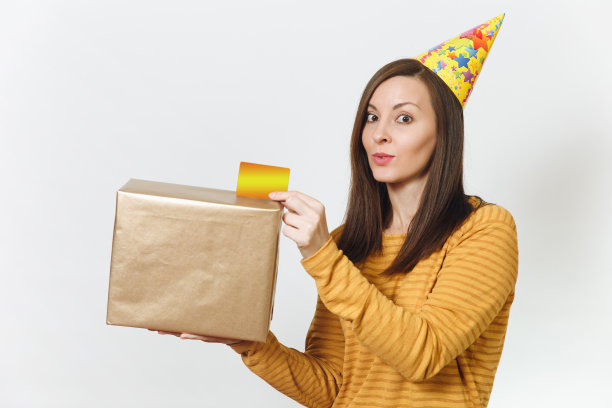 美丽的白人有趣的年轻快乐的女人在黄色的衣服，生日派对帽子拿着金礼盒与礼物和信用卡，庆祝节日在白色背景孤立的广告图片下载