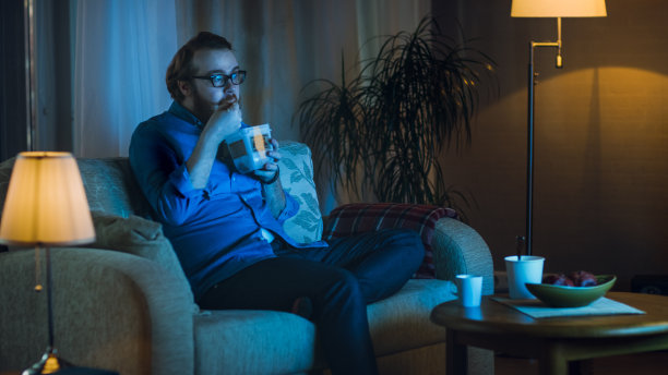 《晚间秀》中，男人坐在客厅的沙发上。他在吃食物盒里的亚洲食物和看电视节目。素材图片