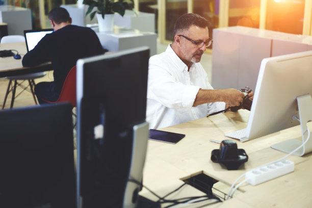 高级成熟的男性业余爱好者或摄影师，在复古相机上设置眼镜模式，在工作后与电脑坐在桌面，在共同的工作空间，使用现代技术制作很酷的照片图片下载