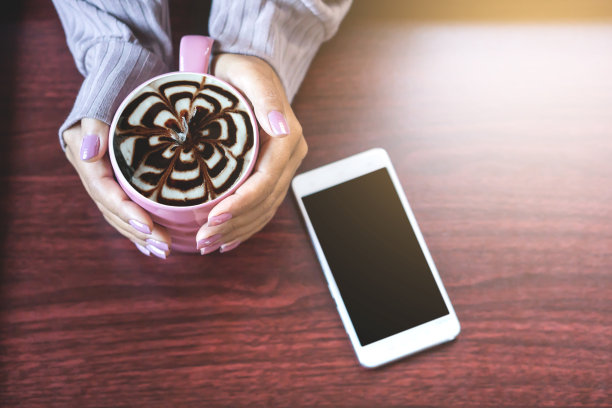 一个女人在木桌上拿着一杯热咖啡玛奇朵，上面涂着奶油和巧克力，还有智能手机，图片下载