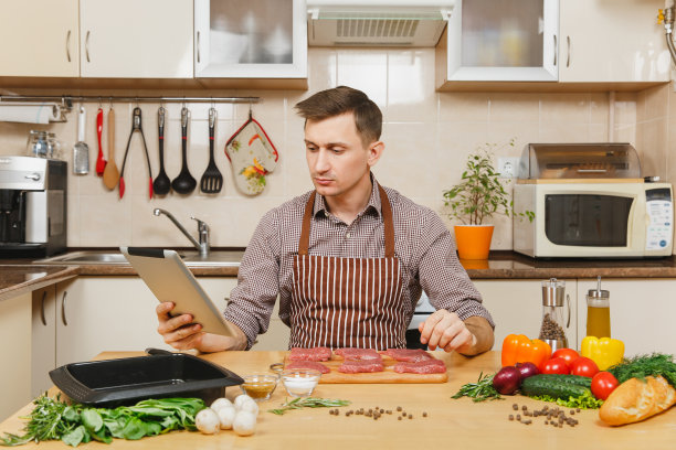 年轻的欧洲男人穿着围裙坐在蔬菜桌前，寻找食谱，在家烹饪，准备猪肉，牛肉或羊肉，在明亮的厨房里，满是高档厨具。图片下载