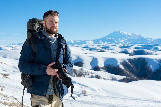 一位留着胡子的时尚摄影师背着背包，戴着太阳镜，手里拿着一台单反相机坐在那里图片下载