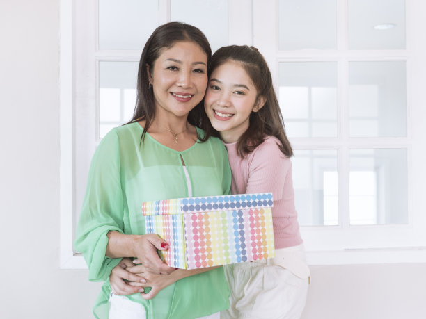 母亲拿着十几岁的女儿送的礼盒，女儿抱着妈妈的窗户背景，爱和关心妈妈的概念。图片下载