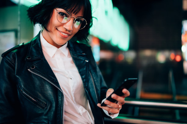 微笑的黑发女人穿着时髦的皮夹克在手机上发信息的肖像，迷人的快乐的时髦女孩戴着眼镜通过手机在晚上休息的时候上传照片到社交网络图片下载