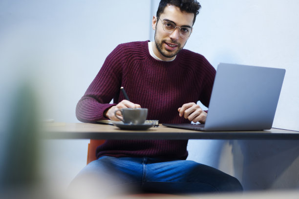 这张照片拍摄的是一名戴着时尚眼镜的大胡子学生，坐在笔记本电脑前，喝着美味的咖啡，看着学校内部的摄像头，一边写作文，一边准备即将到来的考试图片下载