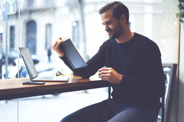 聪明熟练的学生在空闲时间在餐厅内部阅读有趣的书。一个聪明的嬉皮士穿着黑色的运动衫，坐在咖啡店里用笔记本电脑欣赏文学图片下载