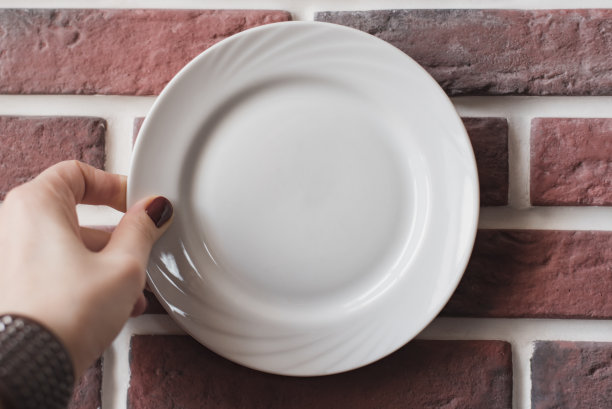 一个戴着棕色皮制手镯的女人的手拿着一个白瓷盘，靠在红砖墙上图片下载