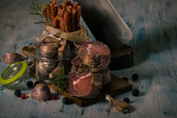 层叠的烤干意大利野猪肉的水平照片。肉放在老式的菜板上，蓝色的木桌上，还有香肠、大蒜、黑刺和大菜刀。图片下载