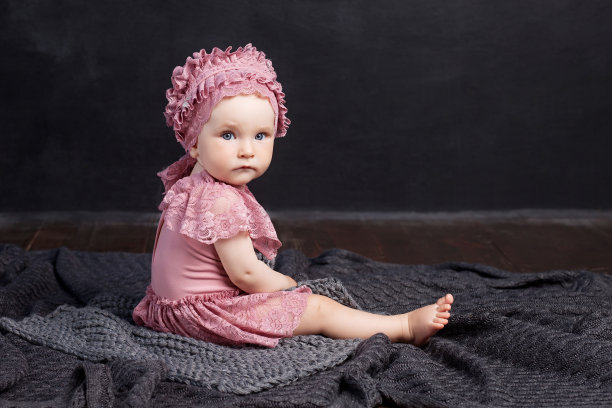 那个女孩坐在地板上的一件灰色编织的格子布上。女孩看着镜头。认真的看。黑色背景。图片下载