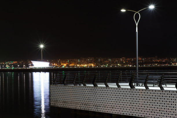 城市的灯光。城市建筑夜间照明。前景是路灯的码头，背景是海堤和灯塔。图片下载