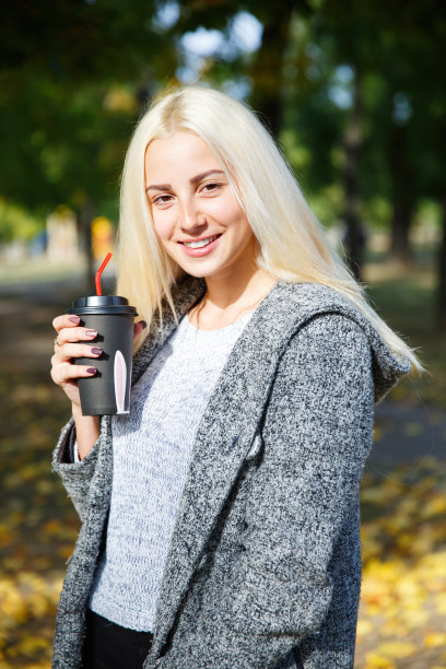 白天，一个身穿灰色夹克的金发女郎在街上端着一杯咖啡。图片下载