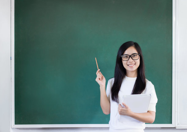 一位亚洲妇女戴着眼镜，手里拿着书和铅笔站在绿色的黑板前图片下载