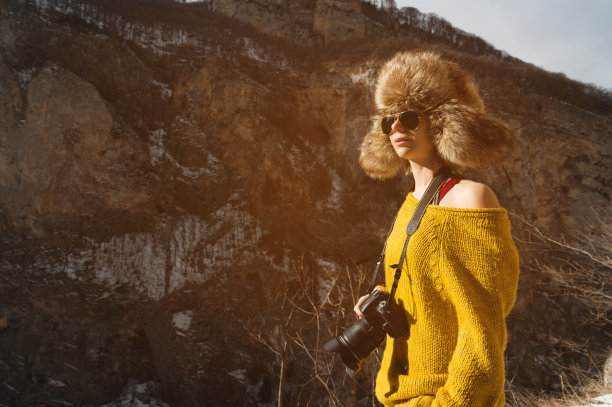 一位戴着太阳镜、戴着一顶大皮草帽、身穿黄色针织毛衣的女摄影师手里拿着一台单反相机，站在峡谷里高高的岩石的背景下图片下载