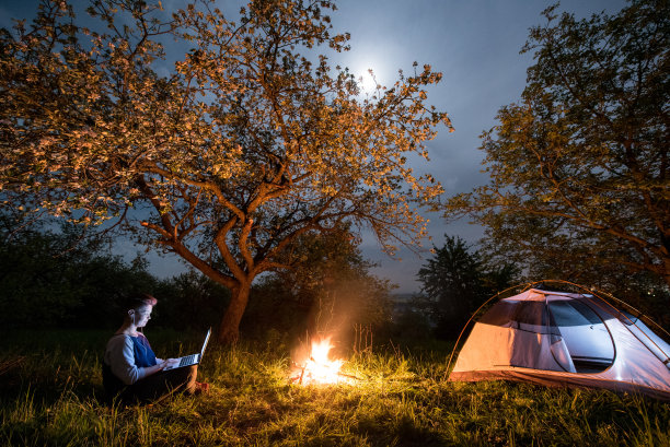 女游客晚上在露营时使用笔记本电脑。女人坐在篝火和帐篷旁，在树下，在有月亮的夜空下图片下载