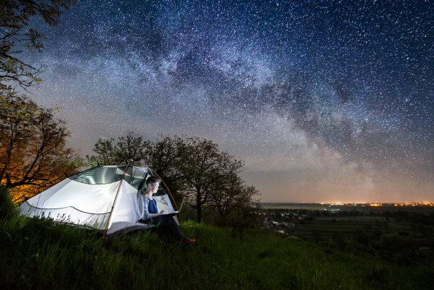 一个晚上在露营用笔记本电脑的女人。女人坐在帐篷里，在美丽的夜空下满是星星和银河。在背景上发光的村庄。长exprose图片下载