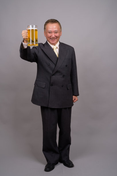 在灰色的背景下，一个成熟的亚洲商人拿着一杯啤酒图片下载