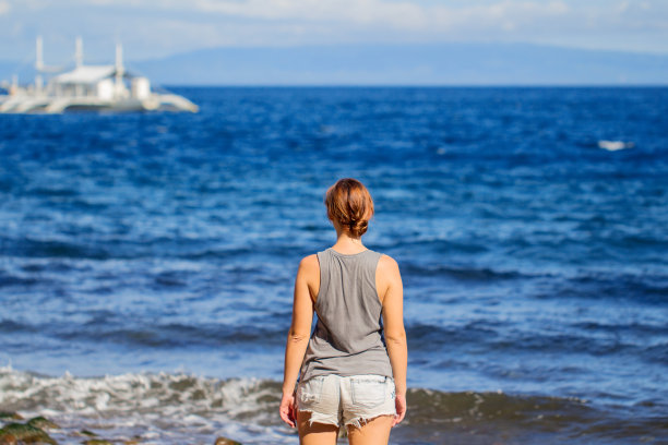 在海滩上穿着衬衫和短裤的女人。年轻女孩看游船的充满活力的照片。图片下载