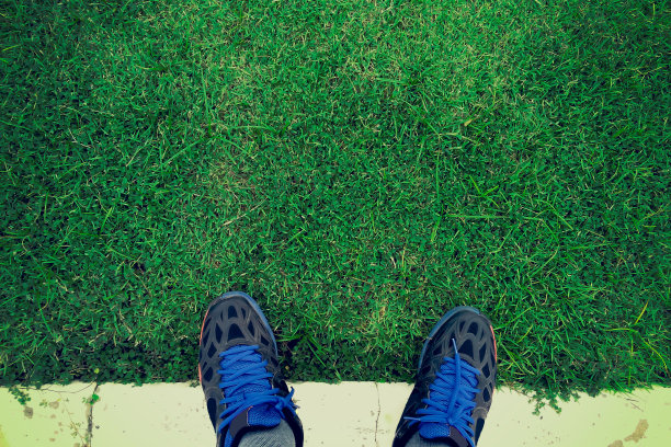 在水泥地板和绿色草地上穿着运动鞋的男人脚的自拍。图片下载