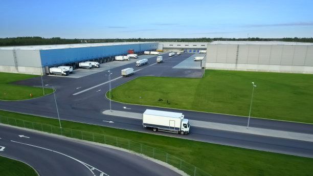 航拍一辆挂着半挂车的卡车离开工业仓库/仓库大楼/装卸区，那里有许多卡车在装卸货物。图片下载