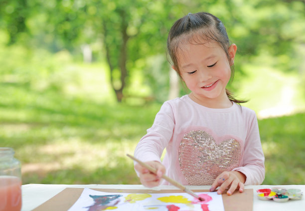 快乐的孩子坐在桌子上，在绿色的花园里画画纸。图片下载