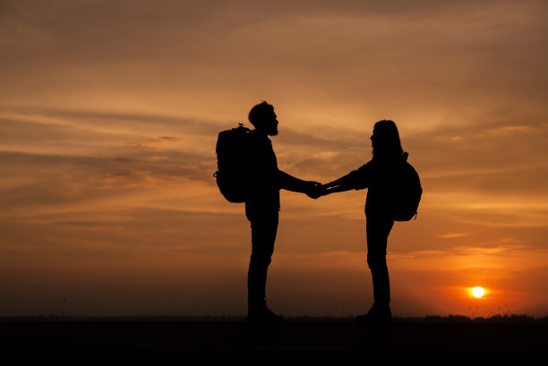 两个背包客(男人和女人)的剪影正在旅行。背景图片是泰国的日落。男人和女人手牵着手站着。图片下载