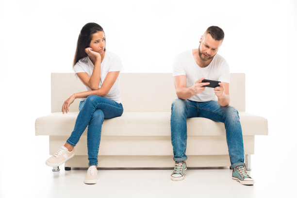 照片中，一名年轻男子正在用智能手机玩电子游戏，他悲伤的女友坐在旁边，孤独地坐在白色屏幕上图片下载