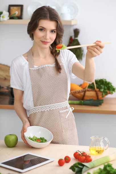 年轻的黑发女子正在厨房里做新鲜的沙拉，并品尝着。家庭主妇手里拿着木勺图片下载