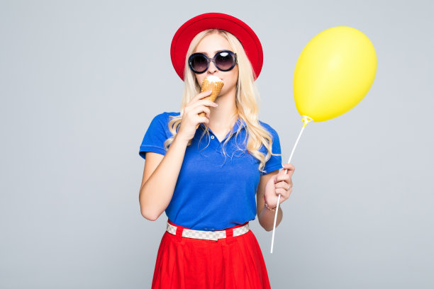时尚快乐微笑的女人与黄色的气球和冰淇淋蛋筒戴草帽和颜色连衣裙在灰色的背景图片下载