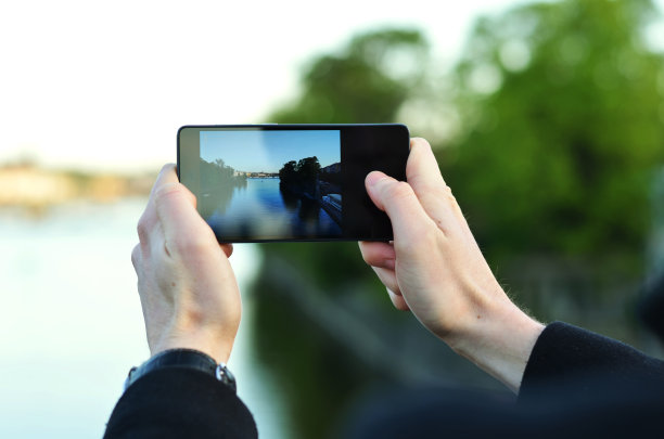 这张照片是一个男人拿着手机在河边拍的照片，背景是模糊的树木和水图片下载