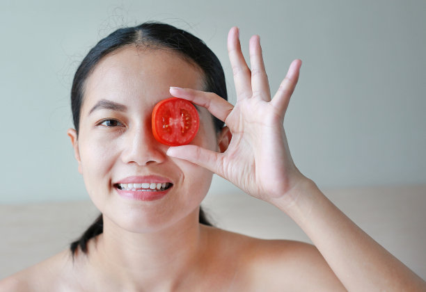 年轻女子拿着西红柿片在她的眼睛，概念皮肤护理。图片下载