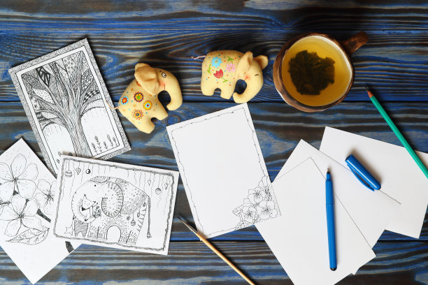 许多明信片的涂鸦风格，画的图片，铅笔，钢笔和陶瓷杯，茶柠檬和两个织物玩具小大象在木制的蓝色背景。手绘，创意，幽默，想法。前视图。铭文。本空间图片下载