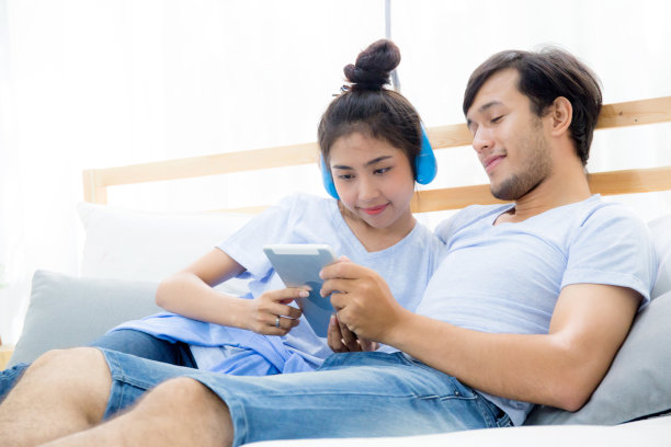 美丽的亚洲年轻夫妇听音乐与平板电脑在床上，爱，约会，年轻夫妇在一起坐在床上使用平板电脑。图片下载