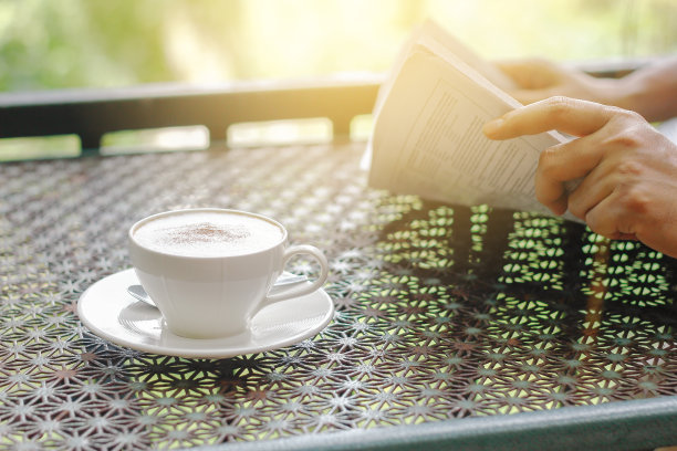股票的照片: 卡布奇诺咖啡杯放在桌子上，背景是一个商人在晨光中读报纸。图片下载