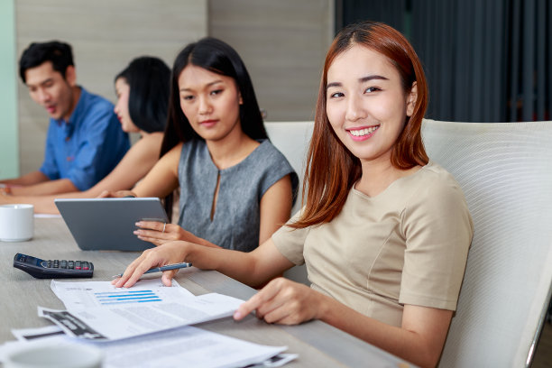 这是一幅年轻的亚洲女商人在会议室里对着镜头微笑的照片，背景是一群工作人员图片下载