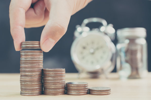 男女手把硬币堆叠起来，以模糊时钟和存钱罐为背景，理财和投资理念素材图片