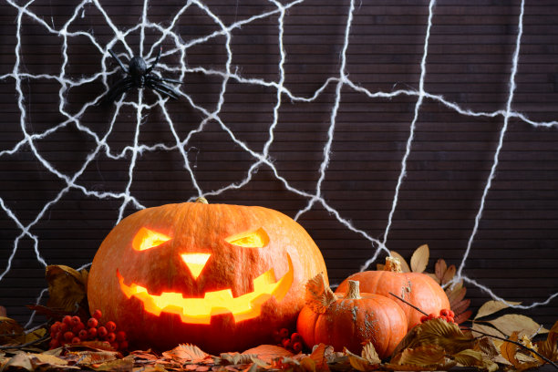 在蜘蛛网的背景上，黄色的树叶中有一张可怕的发光的脸的万圣节南瓜图片下载