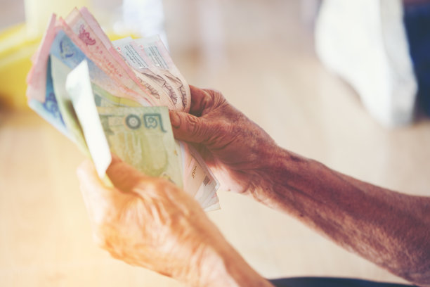 把钞票递给老人用来储蓄的泰国钱。省钱的理念，受益于老年人，灰胡子拿着和展示钱在手里，泰国当地图片下载