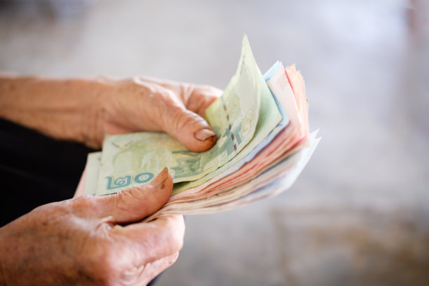 把钞票递给老人用来储蓄的泰国钱。省钱的理念，受益于老年人，灰胡子拿着和展示钱在手里，泰国当地图片下载